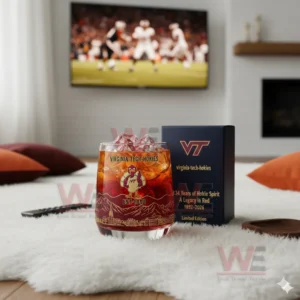 Alt text: A whiskey glass with the Virginia Tech Hokies logo and a commemorative anniversary box, with a football game playing on the TV in the background.