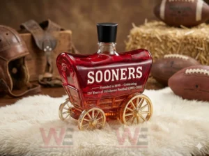 130 Years Of Oklahoma Football 1895-2025 Whiskey Bottle 7 Red wagon style bottle with gold wheels and black stopper placed in a rustic football themed setting!