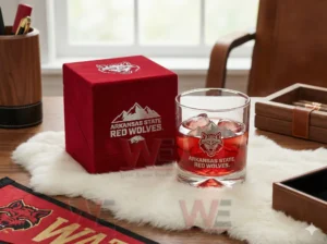 Arkansas State Red Wolves Whiskey Glass 5 A glass containing a red beverage with ice sitting next to a red box on a fur mat with Arkansas State Red Wolves branding visible!