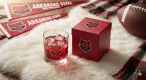 Arkansas State Red Wolves Whiskey Glass 7 A glass containing a red beverage with ice displayed beside a decorated red box with the Arkansas State Red Wolves logo in a cozy sports setting!