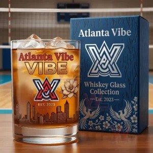 A close up image of an Atlanta Vibe themed whiskey glass containing an amber beverage with ice displayed beside its decorative blue packaging on a wooden surface!