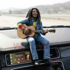 Resin figure of a dreadlocked musician playing an acoustic guitar sitting on a car dashboard above a screen displaying reggae music while driving on a sunny highway!