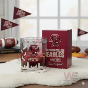 A clear view of a Boston College Eagles whiskey glass featuring feather motifs skyline engraving and red accents displayed beside its branded box with fan decor in the background!