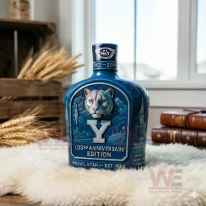BYU 103th Anniversary Edition Whiskey Bottle 5 A blue decorative bottle with a cougar illustration standing on a white furry surface next to wheat and vintage books.