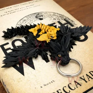Black dragon keychain with yellow eyes and a small golden dragon on its back lying on a Fourth Wing book cover with a metal key ring attached.