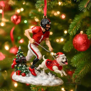 A football player in a red Georgia Bulldogs uniform runs on a snowy ornament base with a bulldog mascot in a red holiday outfit beside a small Christmas tree!