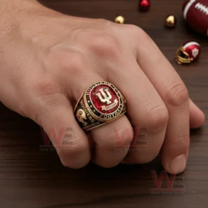 Hand wearing an Indiana Hoosiers football ring with red gemstone style center raised IU logo Indiana banner and Hoosiers Football wording shown on a wooden surface with a football and small decorative items nearby!
