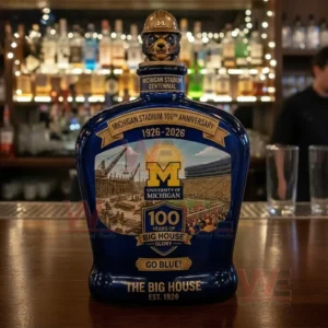 Blue University of Michigan Big House centennial decanter with a wolverine hard hat topper and a detailed anniversary label displayed on a bar counter with a blurred liquor shelf background.