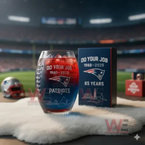 New England Patriots 65th Anniversary Football Whiskey Glass 5 A football shaped glass holding a red and blue drink beside a matching anniversary box on a white fur surface in a stadium environment!