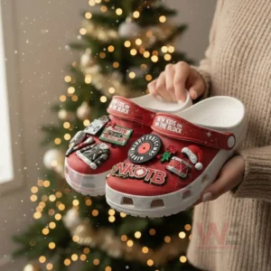 Hands holding red New Kids On The Block clogs decorated with music and Christmas themed charms near a Christmas tree.