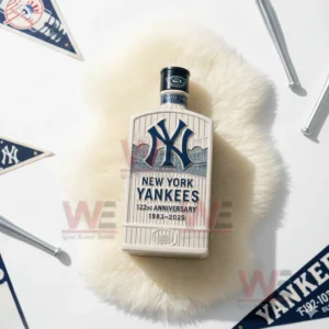 A cream Yankees anniversary bottle with blue logo placed on white fur with baseball pennants and small bats nearby.