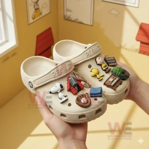 Hands holding beige Peanuts themed clogs decorated with Snoopy Woodstock Charlie Brown and miniature items in a cartoon inspired setting!