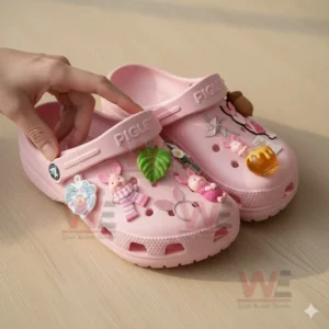 Pink Piglet clogs held by a hand decorated with charms shaped like Piglet figures flowers a leaf a honey pot and a pinwheel.