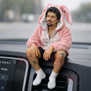 A small figurine of a man wearing a pink bunny hoodie and white socks sitting on a car dashboard with a blurred street view behind!