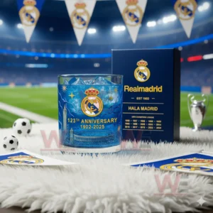 A blue Real Madrid anniversary glass displayed with soccer themed items under stadium lighting.