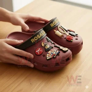 Red Rosalia clogs held by hands decorated with charms including a microphone guitar fan butterfly rose candle and chain.