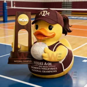 Rubber duck wearing Texas A and M cap and uniform holding a volleyball beside an NCAA trophy on an indoor volleyball court.