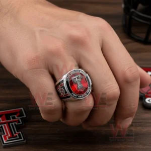 Texas Tech Red Raiders Fight Riders Fight Ring 4 Hand wearing a Texas Tech Red Raiders ring with red gemstone style center raised double T logo and Texas Tech Red Raiders Fight Raiders Fight text on a wooden surface with team themed items nearby!
