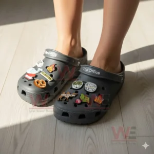 Feet wearing gray decorative clogs adorned with charms referencing an office comedy series shown in bright natural light!