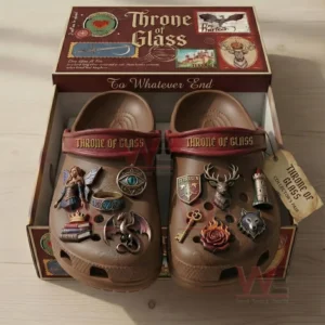 Open shoebox showing Throne of Glass themed brown clogs with maroon straps and gold lettering, decorated with fantasy charms including fairy dragon eye ring books crown crest deer key rose lighthouse and wolf.