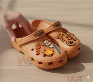 Orange Tigger clogs held by a hand decorated with charms shaped like Tigger faces carrots springs leaves and paw prints.