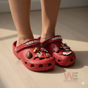 A person wearing red Travis Kelce themed clogs decorated with various football charms on a sunlit wooden floor!