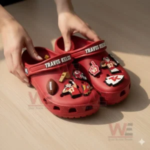 Hands holding red Travis Kelce clogs covered with detailed football related charms on a wooden surface!