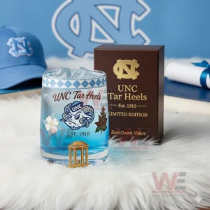 UNC North Carolina Tar Heels Esse Quam Videri Whiskey Glass 3 UNC Tar Heels whiskey glass with blue beverage and ice next to a brown collector box set in a comfortable room decorated with team themed items!