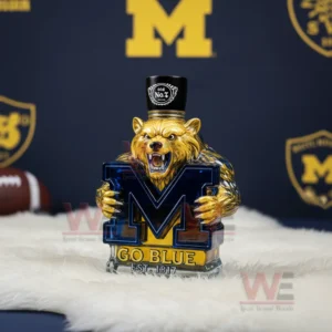 A bear statue holding a blue M emblem with a black top piece standing on a white furry surface in front of Michigan themed decor!