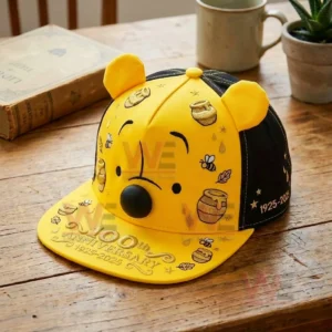 A yellow and black anniversary cap with a bear face honey themed illustrations and playful details displayed on a wooden table with a book and coffee cup in the background!