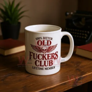 White ceramic coffee mug with a distressed red skull and wings graphic and bold text about being an old bitter club lifetime member on a wooden table with blurred books and a plant in the background!