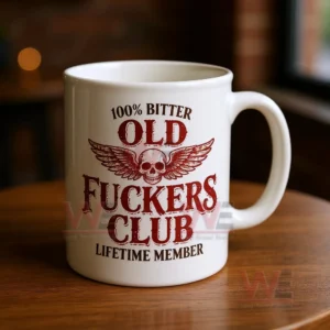 Close up of a white ceramic mug with a distressed red skull and wings design and bold text about being an old bitter club lifetime member on a wooden table with a warm blurred background!