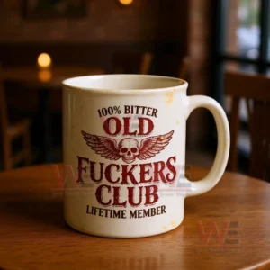 Cream colored ceramic mug with slight stains and a distressed red skull and wings graphic and bold text about being an old bitter club lifetime member on a wooden table with a warm blurred cafe background!