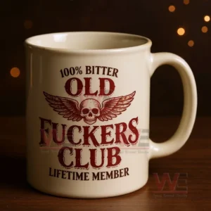 White ceramic mug with a distressed red skull and wings design and bold text about being an old bitter club lifetime member on a wooden surface with warm golden bokeh lights in the background!