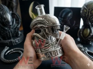 A person holding a silver alien xenomorph glass bong with clear neck and angled bowl with alien themed items and posters behind!
