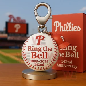 Phillies baseball keychain with red P logo Ring the Bell text 1883 to 2025 142nd and an orange anniversary box on a tabletop in a stadium setting.