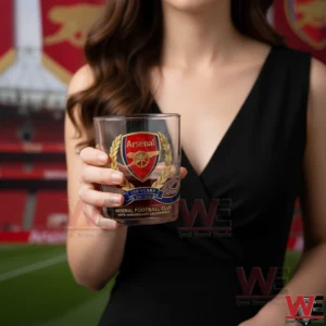 Arsenal 140th Anniversary whiskey glass 6 A close view of a woman holding an Arsenal anniversary whiskey glass with gold and red design elements set against a football stadium backdrop!