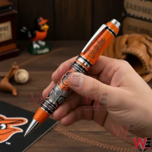 Hand holding an orange Baltimore Orioles anniversary pen with silver accents and engraved team logo in a baseball themed setting.