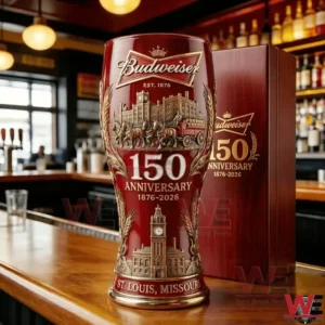 A red Budweiser one hundred fifty anniversary beer glass standing on a wooden bar with a pub interior and bottle shelves behind it!
