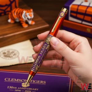Clemson Tigers 130th Anniversary 1896-2026 Pen 3 Hand holding an orange and gold Clemson Tigers commemorative ballpoint pen with purple accents tiger artwork and anniversary text against a Clemson Tigers themed background.
