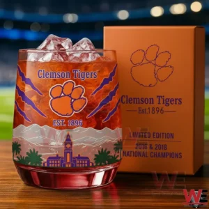 A Clemson Tigers whiskey glass with ice and orange drink featuring the tiger paw logo Clemson Tigers text est 1896 and stadium skyline artwork beside an orange limited edition gift box on a wooden surface