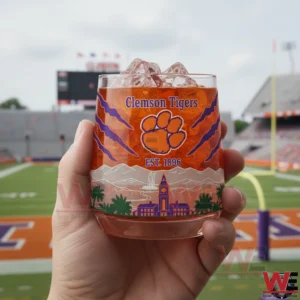 Clemson Tigers 2016 2018 National Champions Whiskey Glass 3 A hand holding a Clemson Tigers whiskey glass with ice and orange drink featuring the tiger paw logo Clemson Tigers text est 1896 and scenic campus artwork in front of a blurred football stadium