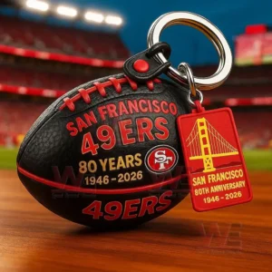 Black football keychain with San Francisco 49ers 80 years anniversary design and Golden Gate Bridge tag on a table with stadium lights behind.