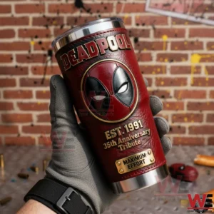 A close view of a Deadpool themed tumbler held in a gloved hand with a brick wall background red leather texture gold mask emblem and strong visual contrast!