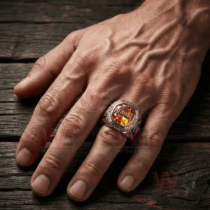 Human hand resting on a wooden surface wearing a large Def Leppard themed ring with orange gemstone face logo text border and detailed metal band design