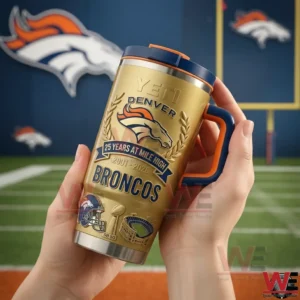 Hands holding a gold Denver Broncos Yeti tumbler with navy lid and orange handle featuring a Twenty Five Years At Mile High Two Thousand One to Two Thousand Twenty Six design in front of a football field background.