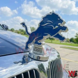 Detroit Lions blue lion hood ornament on a chrome base attached to a cars hood with a bright outdoor sky and parking lot in the background.