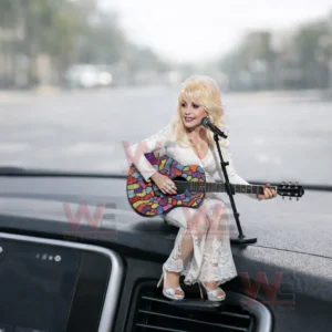 Miniature blonde singer figure in a white dress playing a colorful guitar on a car dashboard with a microphone stand in front.