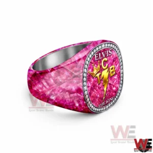 Elvis Presley TCB The King of Rock n Roll Luxury Ring 5 Angled view of a thick pink gemstone textured ring with an oval face reading ELVIS and THE KING featuring a gold TCB crown and lightning bolt emblem surrounded by diamond like stones on a white background!