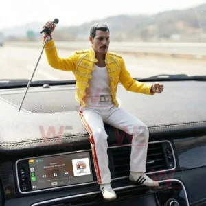 Rock singer figurine in a yellow jacket and white pants holding a microphone while sitting on a car dashboard above a music display with a blurred road background!